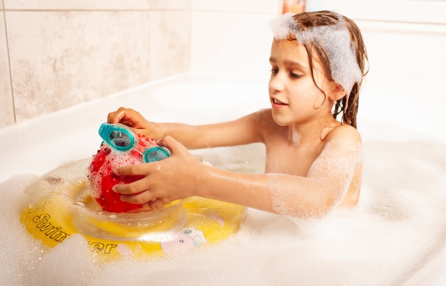 Drôle de petite fille se baigne dans un bain avec de la mousse et joue au ballon de sauvetage