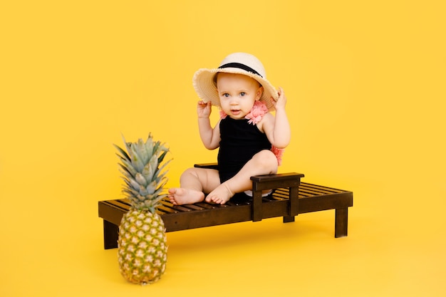 Drôle de petite fille vêtue d'un maillot de bain noir et rose, grand chapeau assis sur une chaise longue en bois avec ananas isolé sur jaune