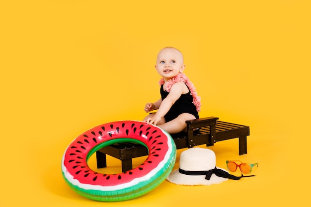 Drôle De Petite Fille Vêtue D'un Maillot De Bain Noir Et Rose, Grand Chapeau Assis Sur Une Chaise Longue En Bois Isolé Sur Fond Jaune. Concept De Vacances D'été