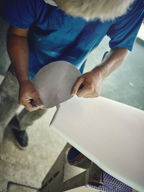 Du haut de la récolte, un maître anonyme polie une planche de surf en bois avec une feuille abrasive dans l'atelier.