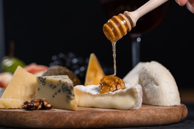 Du miel doux et visqueux est versé sur du camembert aux noix. Différents types de fromages et de fruits se côtoient sur la table.