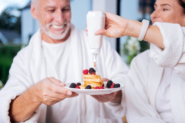 Du miel pour les crêpes. Belle femme portant une montre intelligente blanche à portée de main à l'aide de miel pour les crêpes tout en prenant le petit déjeuner avec son mari