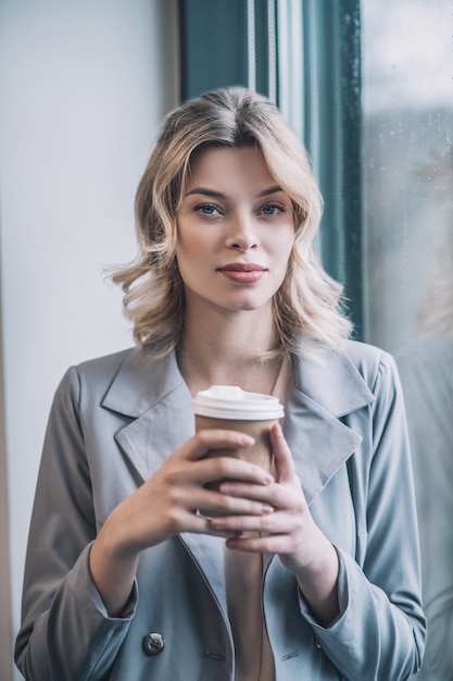Du temps pour toi. Jolie femme blonde d'affaires en veste grise tenant le café dans ses mains repos calme près de la fenêtre au bureau