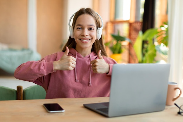 Éducation à distance Adolescente excitée dans un casque montrant les pouces vers le haut et souriant tout en utilisant un ordinateur portable à la maison