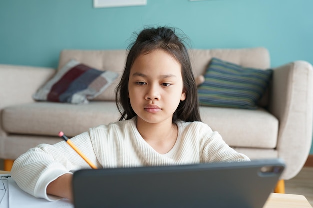 Éducation. Une fille asiatique apprend avec un tuteur en ligne via Internet sur une tablette numérique à la maison le matin. Les enfants d'Asie regardent l'ordinateur et écrivent leurs devoirs. Concept d'apprentissage en ligne à la maison