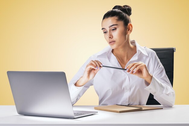 Éducation en ligne et concept d'étude avec une jolie jeune femme assise à une table blanche regardant un écran d'ordinateur portable et portant un crayon dans les mains sur fond de mur orange clair abstrait
