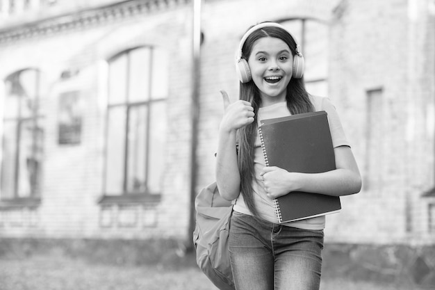 Éducation pour les enfants écoutant de la musique enfance heureuse petite fille porter des écouteurs technologie moderne dans l'éducation fille joyeuse tenir le dossier retour à l'école enfant heureux avec ordinateur portable