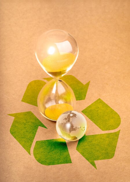 Photo durabilité environnementale temps et responsabilité préserver les ressources concept petit globe du monde en verre près d'un sablier sur un grand symbole de recyclage vert sur un fond de papier brun style vertical