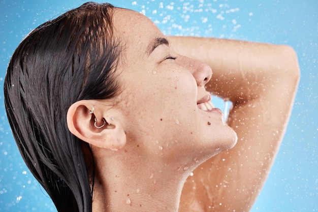 Eau de douche et visage d'une femme nettoyant le corps pour se détendre pour le bien-être sur un fond de studio bleu Soins des cheveux heureux et modèle asiatique se lavant pour le toilettage de l'hygiène et de la santé avec un sourire