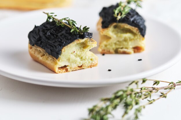 Eclairs à la crème de pistache et glaçage noir. Gâteaux décorés de brins de thym. Dessert sur la table de cuisine blanche.