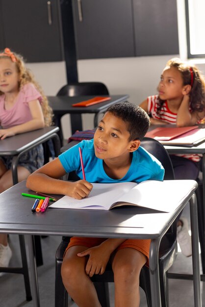 À l'école, un jeune étudiant de race mixte est assis à un bureau dans une salle de classe à écrire.