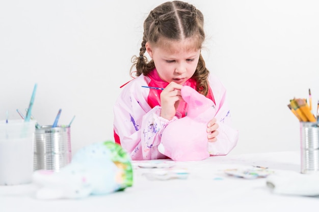 L'école à la maison pendant le confinement lié au COVID-19. Petite fille peignant une figurine de lapin de Pâques en papier mâché avec de la peinture acrylique.