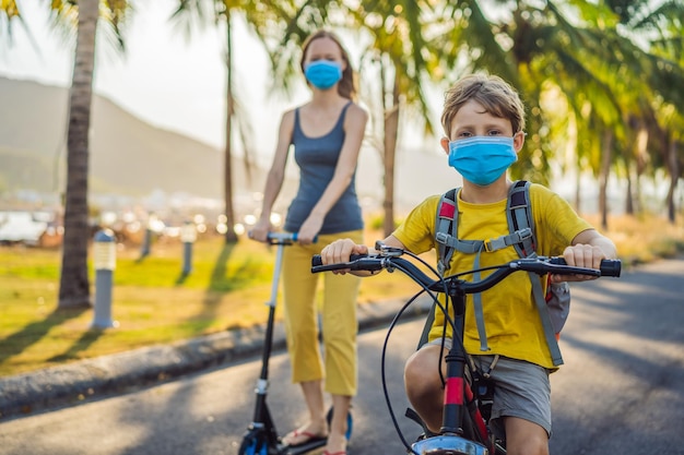 Un écolier actif et sa mère portant un masque médical font du vélo avec un sac à dos par temps ensoleillé Joyeux enfant faisant du vélo sur le chemin de l'école Vous devez aller à l'école avec un masque à cause de l'épidémie de coronavirus