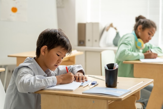 Photo Écolier asiatique prenant des notes dans un cahier par bureau contre son camarade de classe