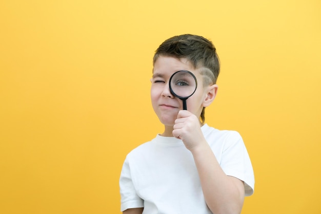 Un écolier curieux et positif dans des vêtements décontractés regarde la caméra à travers une loupe sur fond jaune