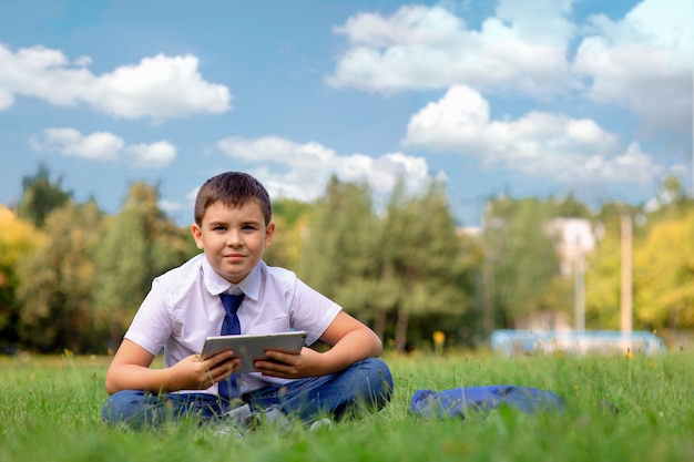 Un écolier dans une chemise blanche et une cravate bleue est assis sur l'herbe verte contre un ciel bleu avec des nuages blancs et tient une tablette.