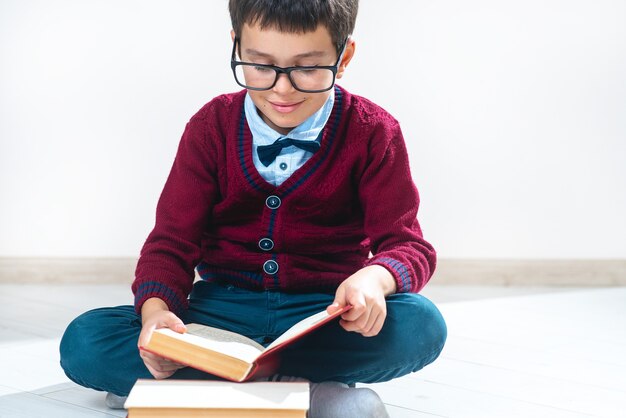 L'écolier dans un pull et des lunettes est assis sur le sol en position du lotus et feuillette un livre