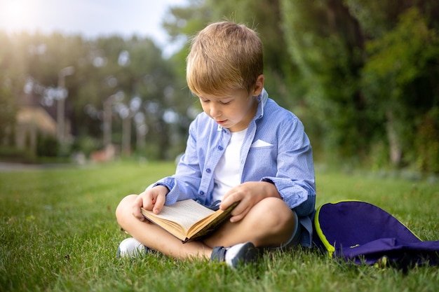 Un écolier est assis sur l'herbe verte dans le parc avec un sac à dos et un livre lisant un livre