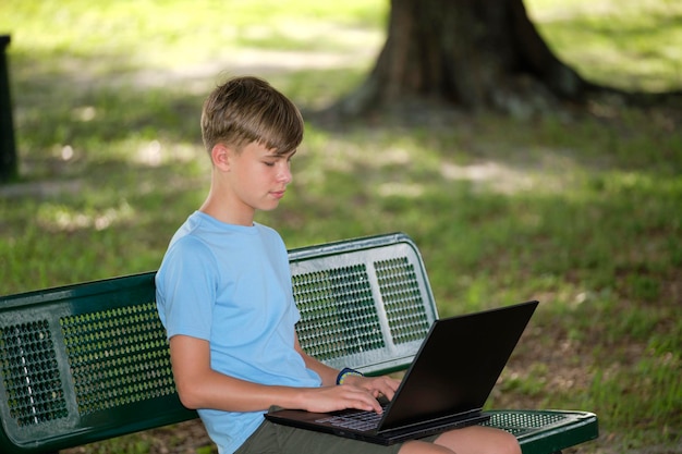 Écolier étudiant sur un ordinateur portable reposant sur un banc de parc à l'extérieur le jour de l'été Enseignement à distance pendant le concept de quarantaine