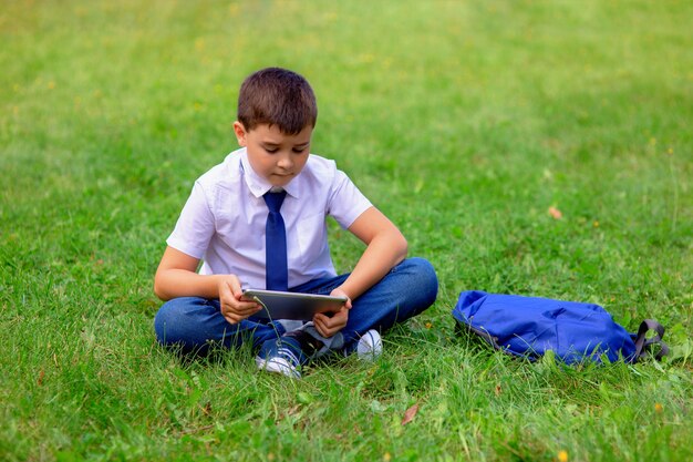 Un écolier heureux dans une chemise blanche s'assied sur l'herbe verte et joue une tablette