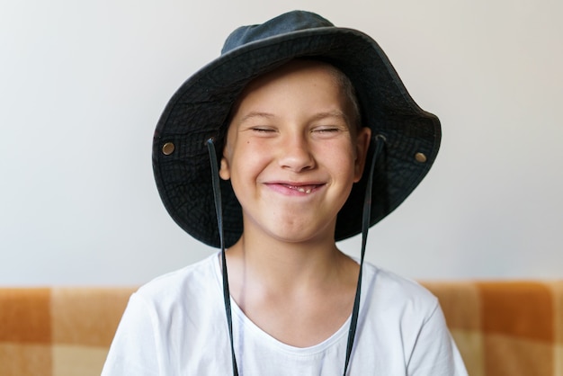 Un écolier joyeux en t-shirt blanc et chapeau noir est assis sur le canapé et fait une grimace amusante...