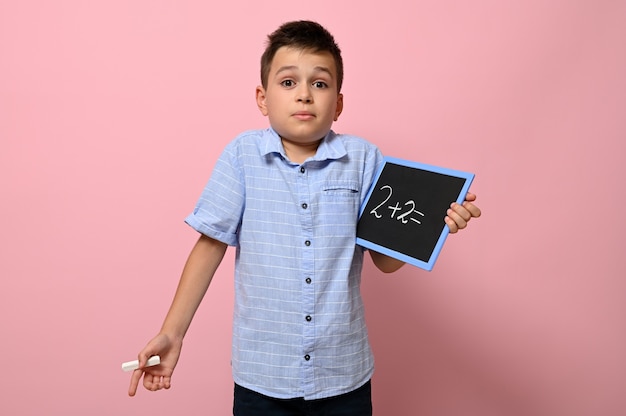 Un écolier mignon tient un tableau noir à la main avec un problème d'arithmétique et avec surprise et questions regarde la caméra en se tenant debout sur un fond rose avec un espace pour le texte. Retour à l'école.