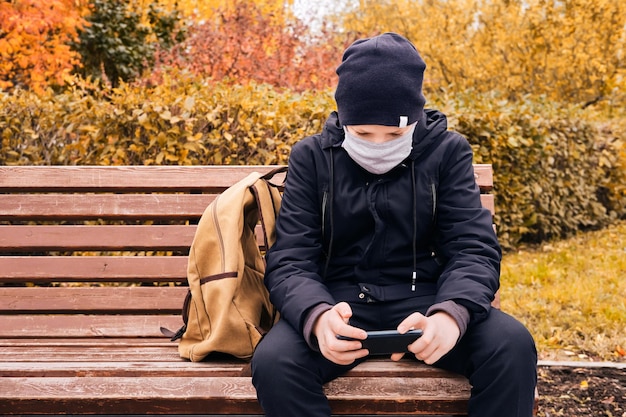 Un écolier portant un masque de protection est assis sur un banc et tient un smartphone Nouvelle normalité