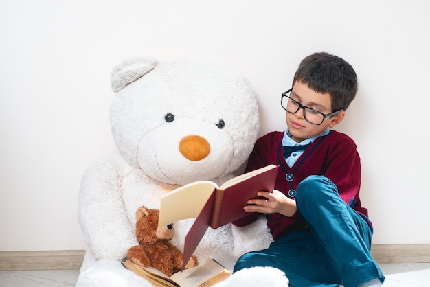 L'écolier en pull et lunettes montre un livre ouvert à un gros ours en peluche