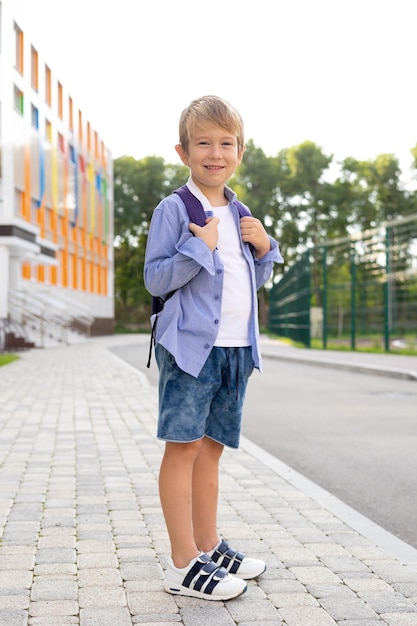 Un écolier se tient avec un sac à dos près de l'école regarde l'école primaire de la caméra