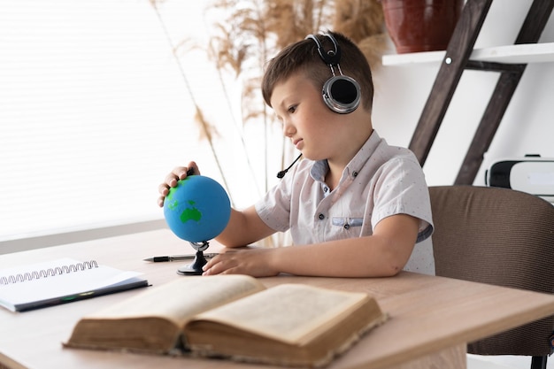 Un écolier triste assis dans des écouteurs à un bureau écoute une leçon de géographie et regarde le globe étudiant le pays et l'océan Casque sans fil et apprentissage en ligne Enseignement à distance