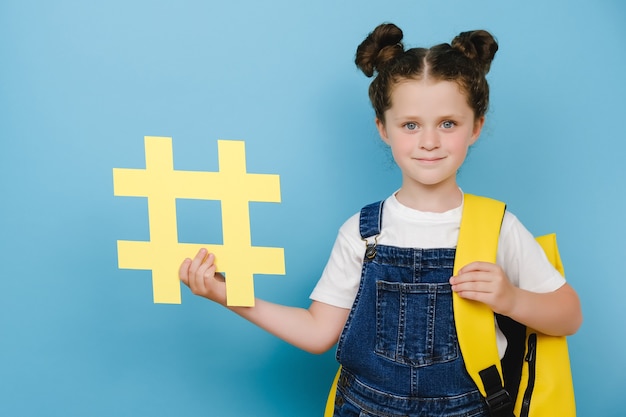 Photo Écolière adorable mignonne avec le sac à dos tenant le grand symbole jaune de hashtag et heureuse regardant la caméra, montrant le signe de hachage, posant sur le fond bleu. concept de publications et d'écoles à la mode sur les réseaux sociaux