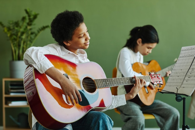 Photo Écolière afro-américaine diligente avec guitare acoustique regardant du papier avec des notes sur le pupitre