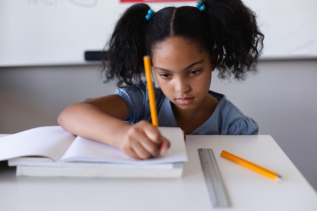 Une écolière binaciale écrit sur un livre à son bureau pendant qu'elle étudie en classe.