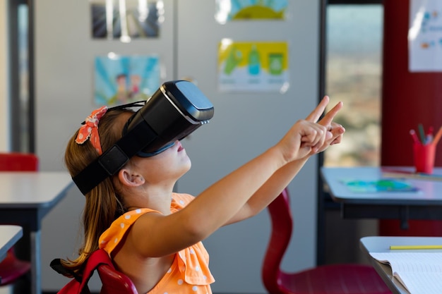 Une écolière caucasienne concentrée assise à son bureau et utilisant un casque VR en classe à l'école primaire