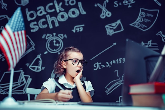 Une écolière est assise à la table avec des livres pour ordinateur portable et un tableau avec des formules scolaires sur fond w...