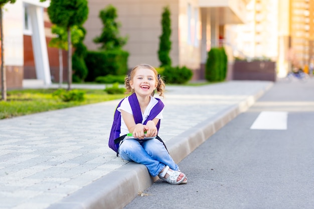 Une écolière heureuse avec un sac à dos et des manuels dans ses mains est assise sur le trottoir à l'école, le concept est de retour à l'école