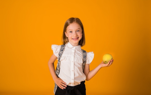 Une écolière joyeuse en blouse blanche et avec un sac à dos tient une pomme verte sur fond jaune avec une copie de l'espace