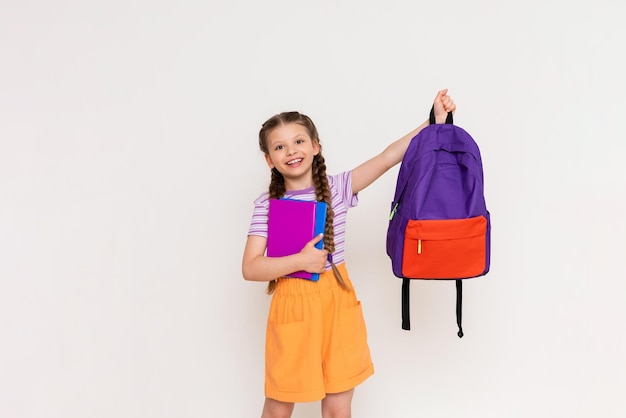 Une écolière avec des livres dans ses mains soulève un sac à dos d'école sur un fond blanc isolé Préparation pour l'année scolaire