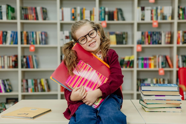 Une écolière à lunettes est assise à un bureau avec une énorme pile de manuels scolaires dans la bibliothèque