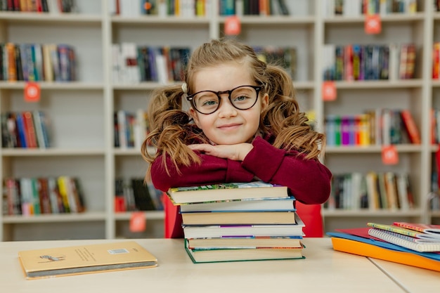 Une écolière à lunettes est assise à un bureau avec une énorme pile de manuels scolaires dans la bibliothèque