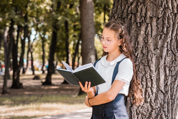Une écolière à lunettes lit un livre près d'un arbre dans le parc