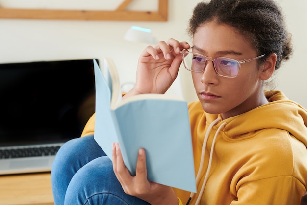 Une écolière métisse sérieuse assise au bureau avec un ordinateur portable et ajustant des lunettes tout en lisant un livre et en se préparant pour la leçon