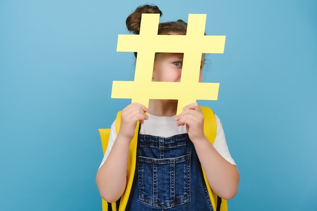 Une écolière mignonne et ludique tient un signe de hashtag en papier jaune, regardant la caméra en s'amusant, porte un sac à dos, posant isolé sur le mur de fond bleu du studio. Concept de publications et d'écoles à la mode sur les réseaux sociaux