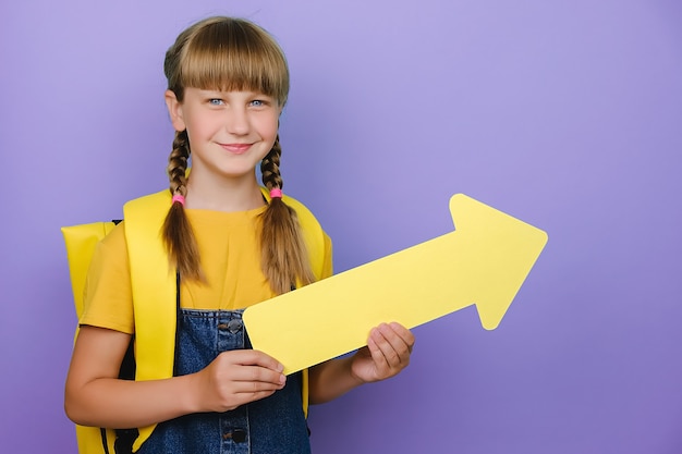 Photo Écolière mignonne souriante montrant de côté avec une flèche jaune, porte un sac à dos, posant isolée sur un mur de fond de couleur violet studio avec espace de copie pour le contenu promotionnel. retour au concept de l'école