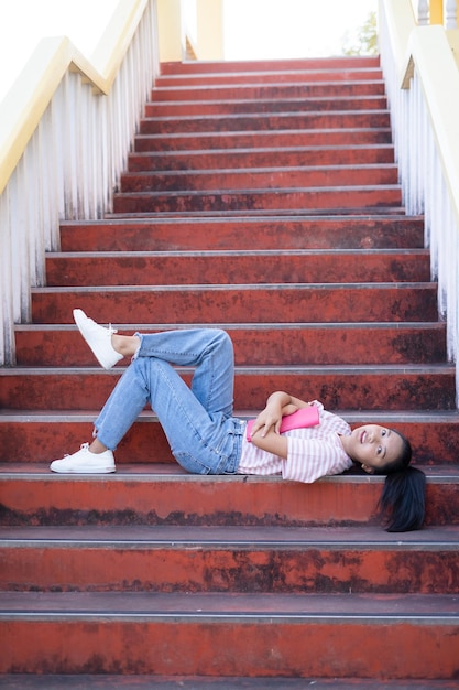 Une écolière porte une chemise rose et un jean tenant et tenant un livre rose dans les escaliers