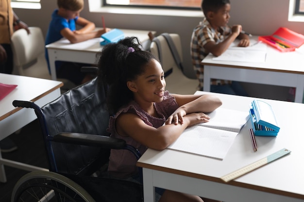 Une écolière primaire biraciale souriante assise sur un fauteuil roulant à un bureau dans la salle de classe.