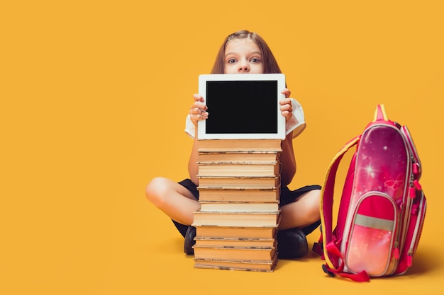 Une écolière regarde par derrière une pile de livres et une tablette regarde l'éducation des enfants de la caméra