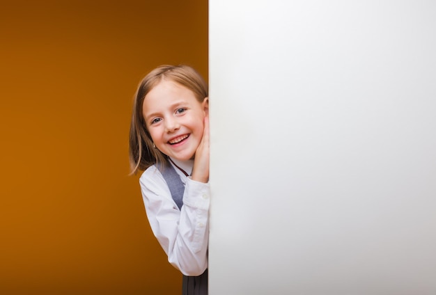 Une écolière souriante se tient derrière un panneau blanc vierge mis en évidence sur un fond jaune Un espace vide pour le texte regarde derrière la bannière