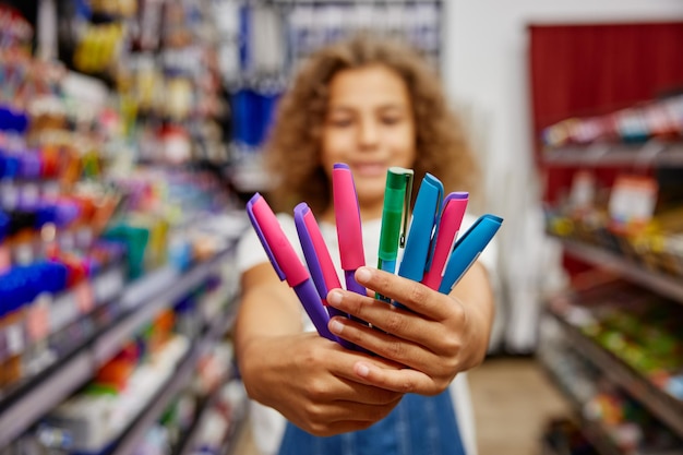 Une écolière tenant une variété de stylos pour écrire debout dans un magasin de papeterie