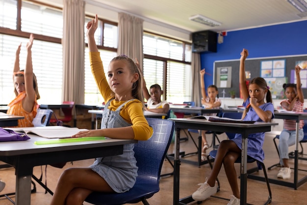 Des écoliers élémentaires divers lèvent la main à des bureaux en classe